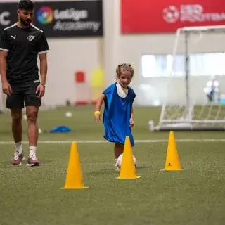 Sports Summer Camps Dubai | A girl dribbles a soccer ball around cones under a coach's supervision