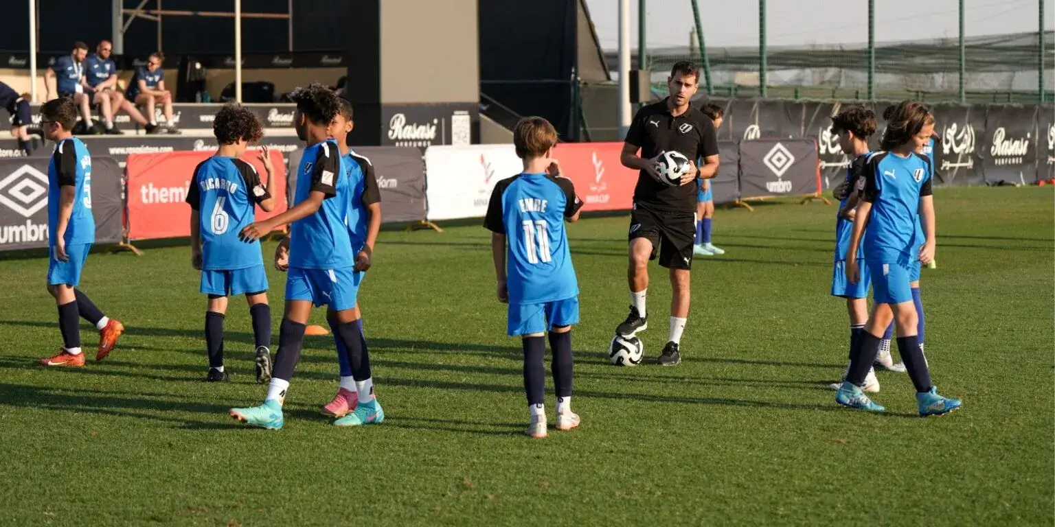 A group of boys in soccer uniforms playing on a grass field with their coach, kicking a ball during a practice match for the Mina Cup 2024.