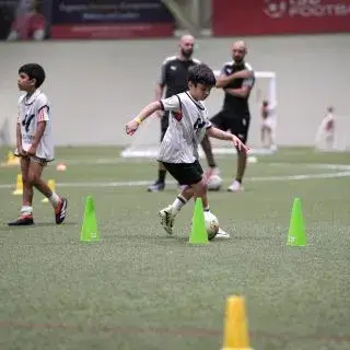 Summer Football Camps |A young boy practicing soccer skills by kicking a ball around cones on a field.